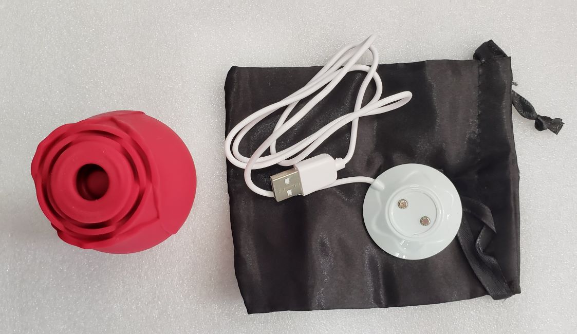 Rose Suction Stimulator: An aerial shot of the rose toy next to a black fabric storage bag with a drawstring closing mechanism and a white charging stand with a USB plug-in attached.