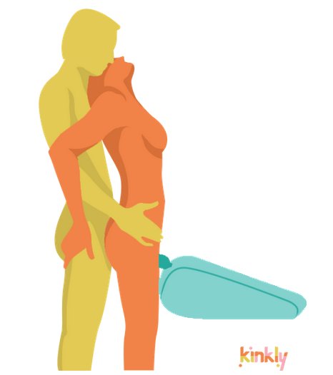 Twin Standing Pleasure Wand Massager Sex Position. A person with a clitoris is standing upright with their clit positioned on top of a wand massager that is being held by a sex shape called the Liberator Axis. The partner of the clitoris owner then comes up from behind to penetrate them. 