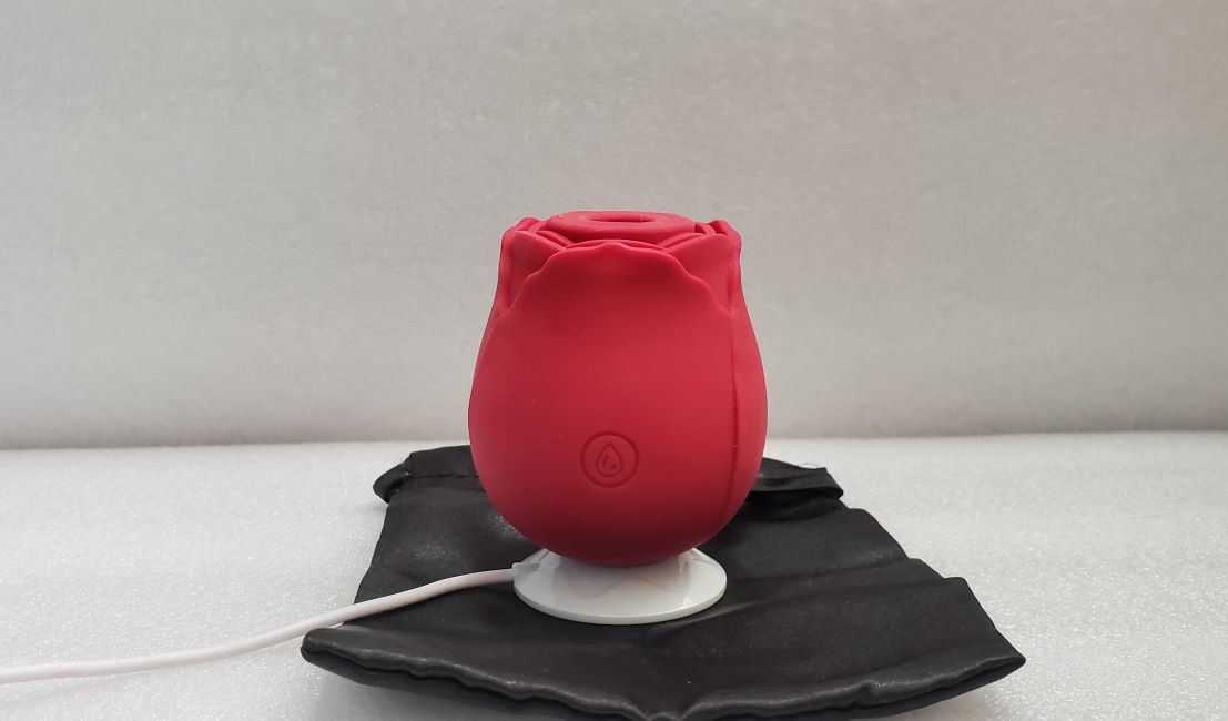 Rose Suction Stimulator: A side view of the rose toy resting on a black piece of fabric against a gray background. The toy is bright red and bulbous with a white stand holding it upright and silicone petals like those of a rose near the top. 