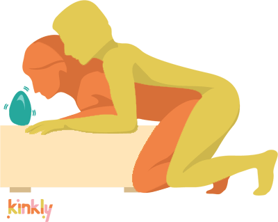 Magic Mountain Sex Position. The receiving partner is leaned over a low ottoman with their forearms on the furniture and their knees on the floor. The penetrating partner copies this position, kneeling right behind the receiver to facilitate sex.