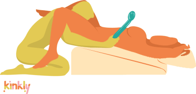 Elevated Oral Sex Position. The receiving partner is laying back on the Liberator Ramp while using the Liberator Wedge as a pillow. The giving partner is kneeling with their face between their partner's legs. The giving partner holds a vibrator in their hand which can be used on the receiving partner. 