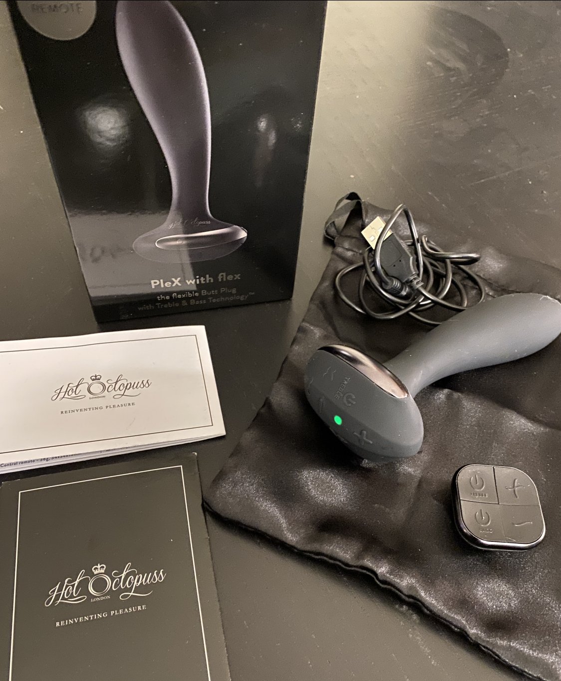 The Hot Octopuss PleX with Flex sits out on a countertop with everything that is included in the packaging. There is the anal vibe, the remote control, the charging cable, the instruction manual, and black drawstring bag.