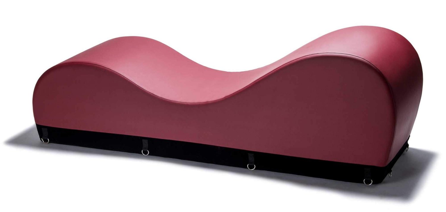 The Liberator Black Label Esse Chaise. A piece of sex furniture with a black base, that features metal loops for attaching bondage equipment, and a foamy burgundy top. The top of the furniture is composed of two rounded sides, of inconsistent heights, and a large divot in the middle.
