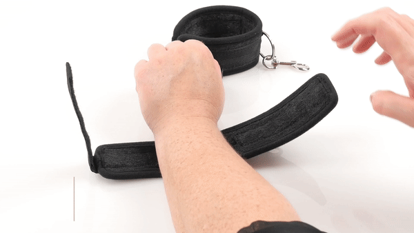 The Sportsheets Soft Cuffs: A pair of black fabric handcuffs with velcro closures and metal attachments to hook the cuffs onto each other or other bondage equipment. A person is shown closing the cuff around their wrist.